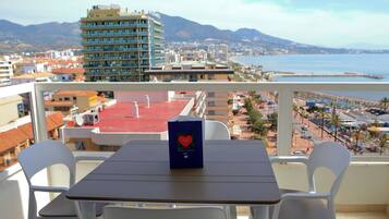 Parvekkeella on pöytä ja tuolit, ja sieltä avautuu näkymä rannikkokaupunkiin, jossa on rakennuksia ja ranta.