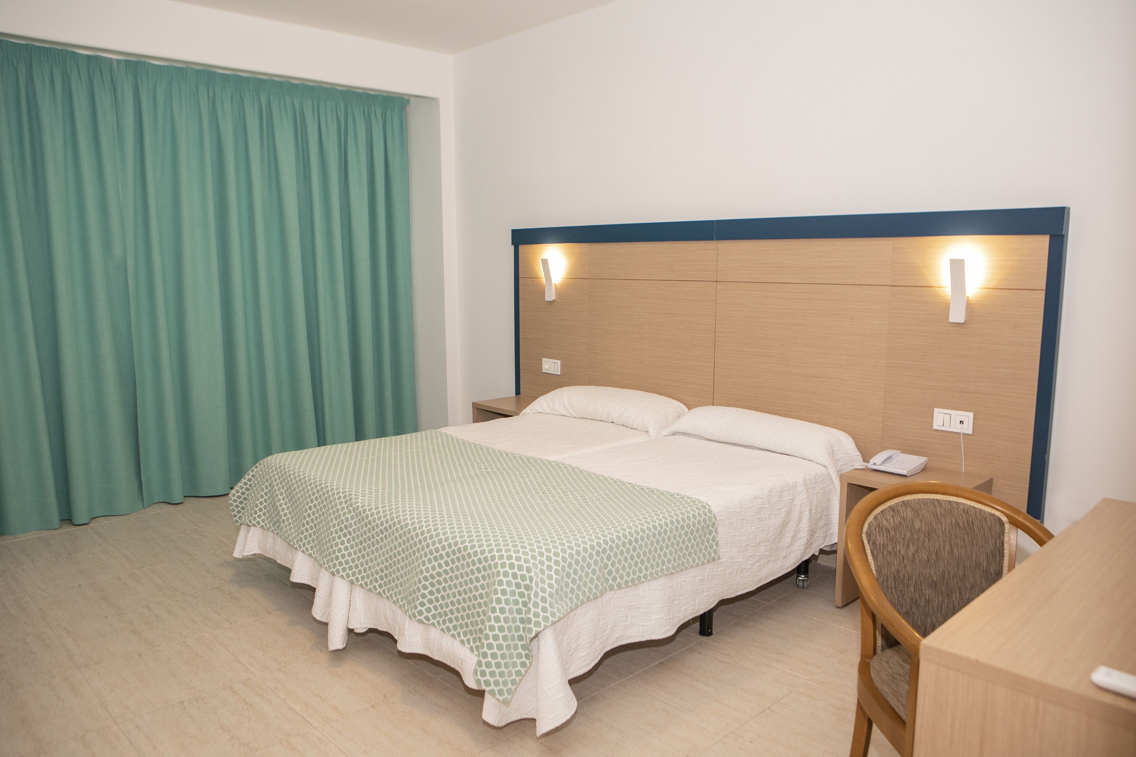 A hotel room with a bed, a desk, and a chair. There is a curtain on the window and a wall-mounted light fixture above the bed.