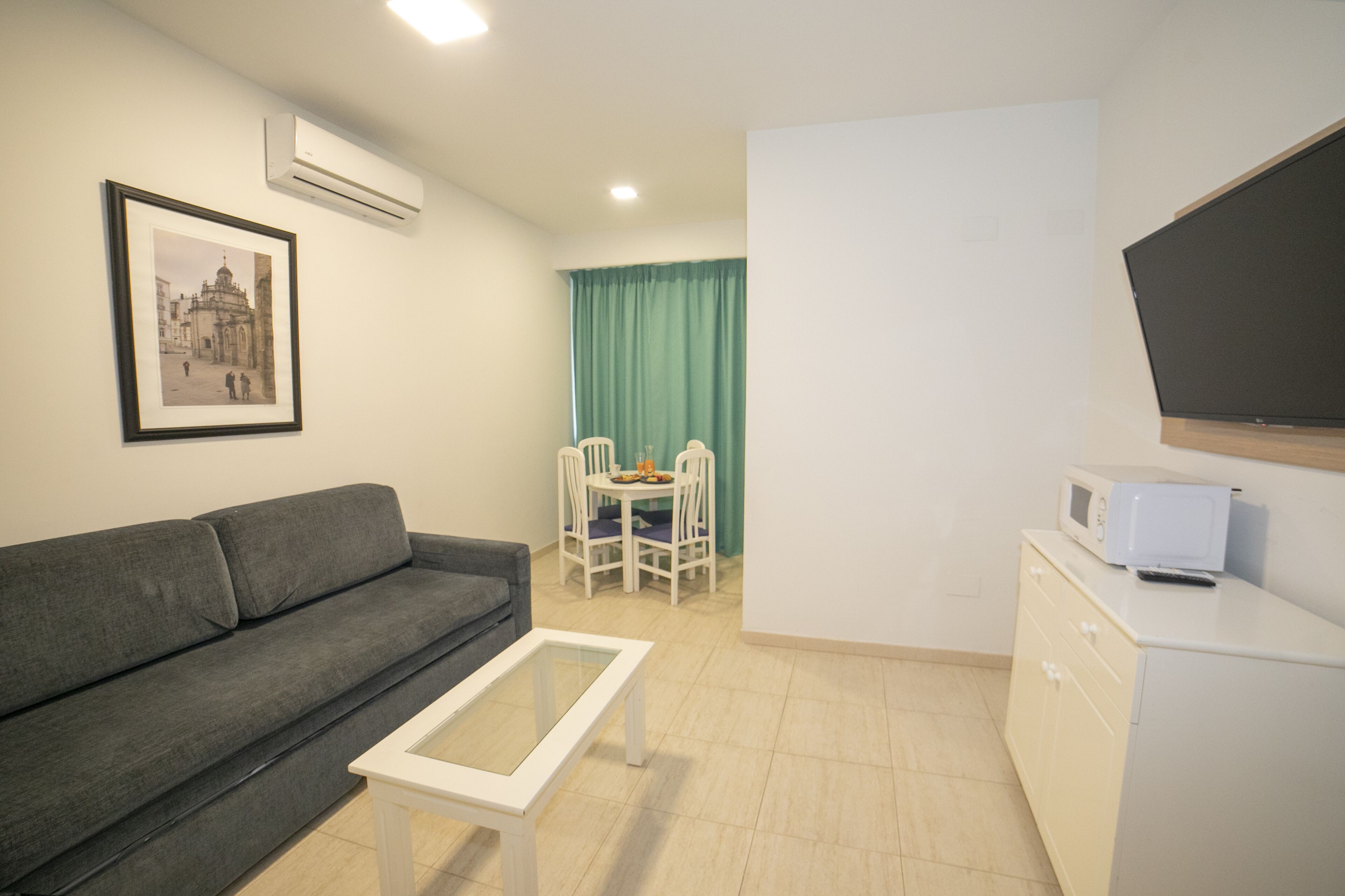 Un espacio habitable compacto con un sofá gris, una mesa de centro blanca, una zona de comedor con mesa y sillas, un microondas en un mueble y un cuadro en la pared.