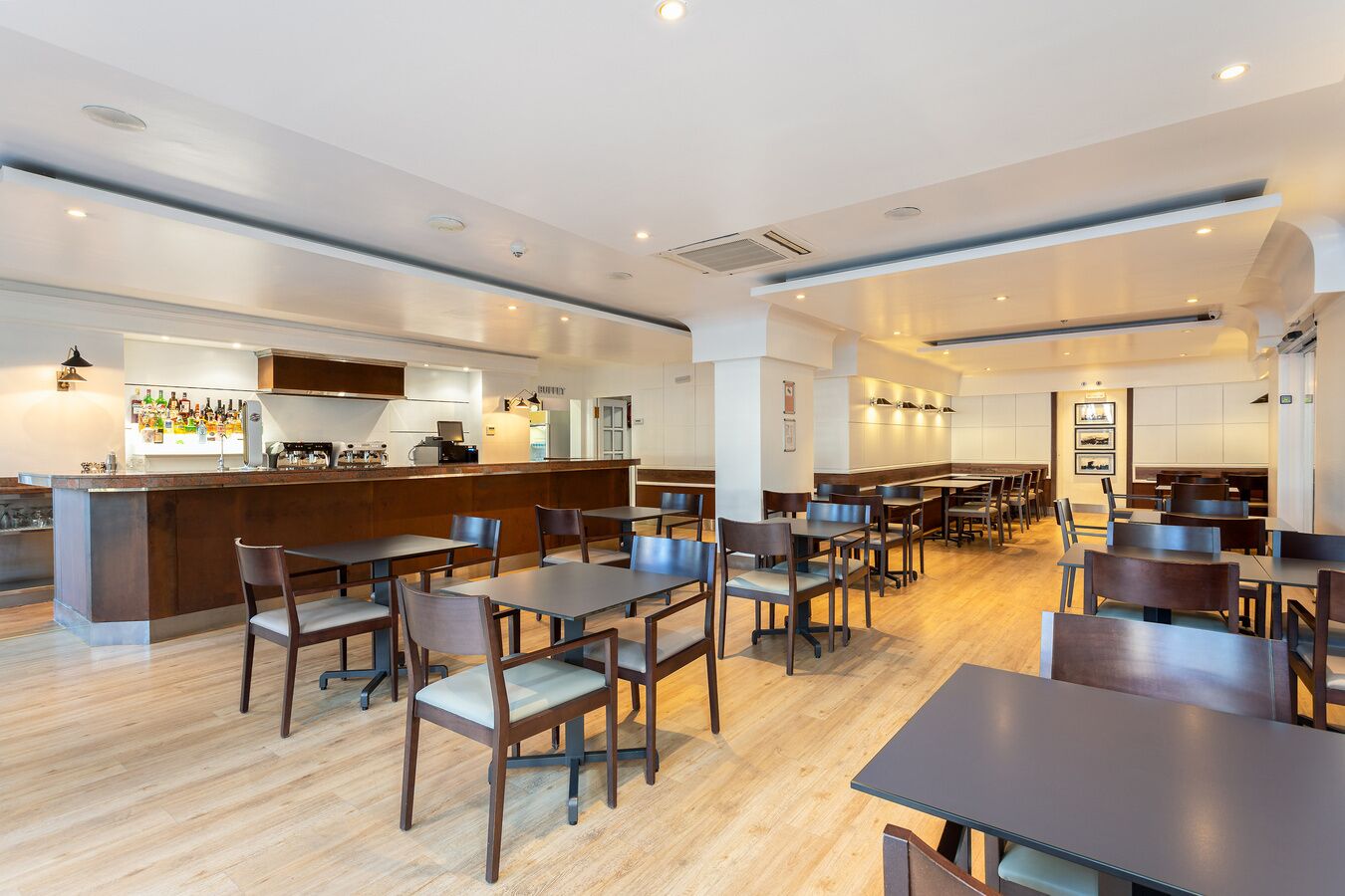 A modern restaurant with wooden tables and chairs, a bar area, and a kitchen in the background.