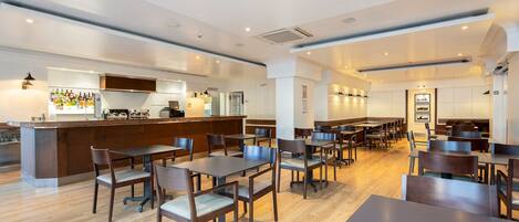 A modern restaurant with wooden tables and chairs, a bar area, and a kitchen in the background.