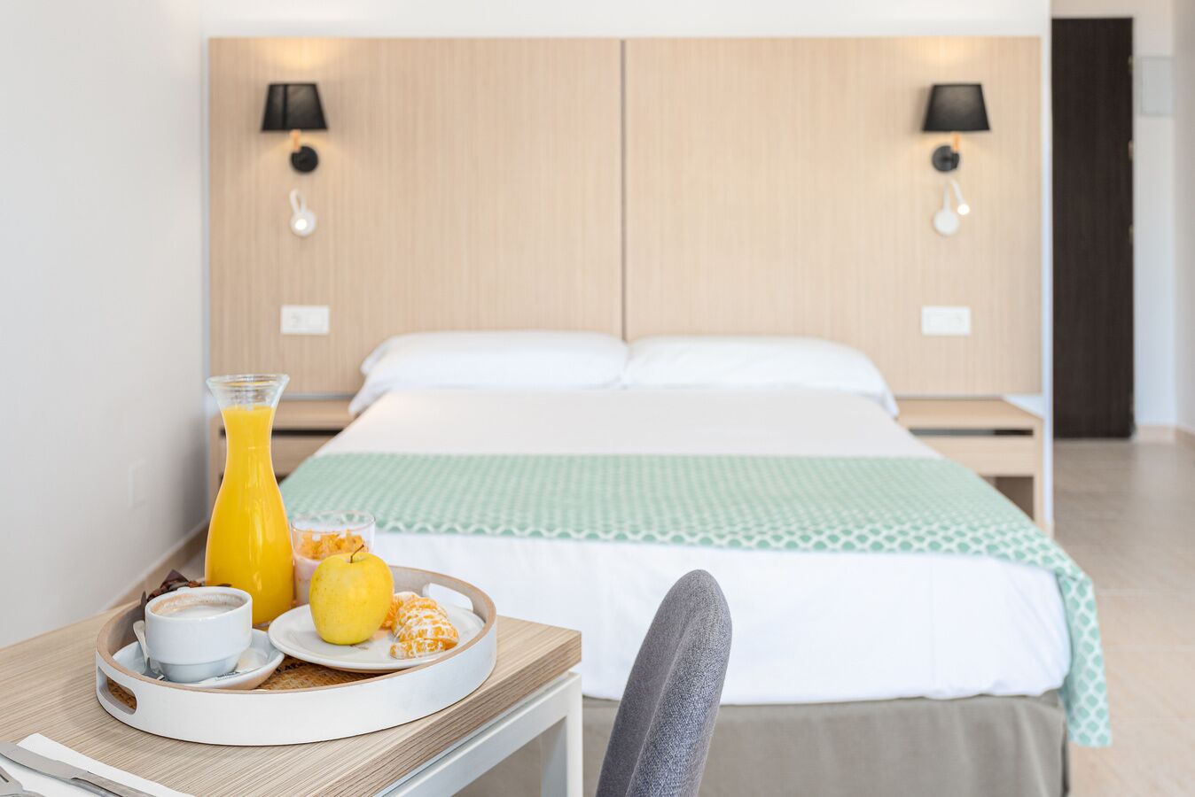 Habitación de hotel con una cama, mesitas de noche, una bandeja de desayuno con jugo de naranja, una taza y un plato con fruta.