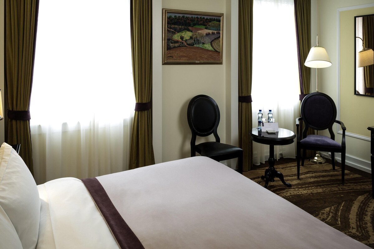 A hotel room with a bed, two chairs, a small table with a lamp, a painting on the wall, and curtains.