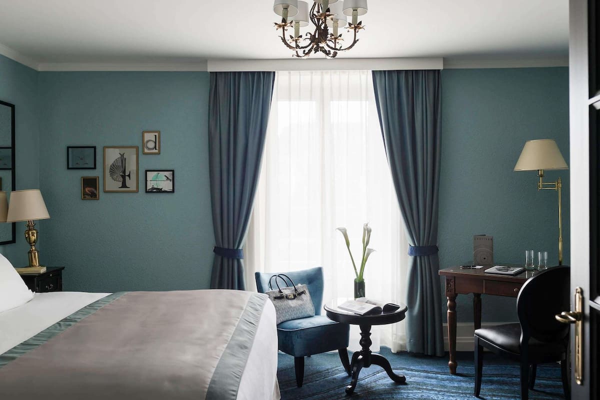 A hotel room with a bed, a chair, a desk, a chandelier, and curtains.