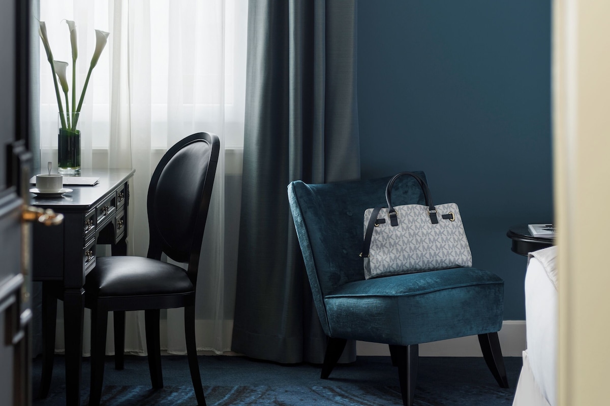 A hotel room with a desk, chair, and a blue velvet armchair with a handbag.