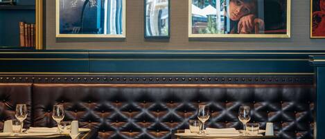 Interior de un restaurante con una zona de asientos en un banco de cuero oscuro, obras de arte en las paredes y una mesa dispuesta con vasos y cubiertos.