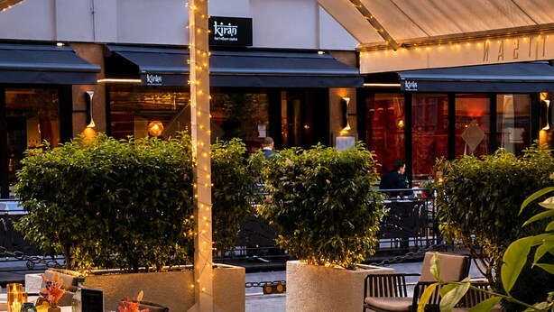 Un área de comedor al aire libre con mesas dispuestas para dos, rodeada de sillas, bajo una estructura cubierta con luces colgantes.