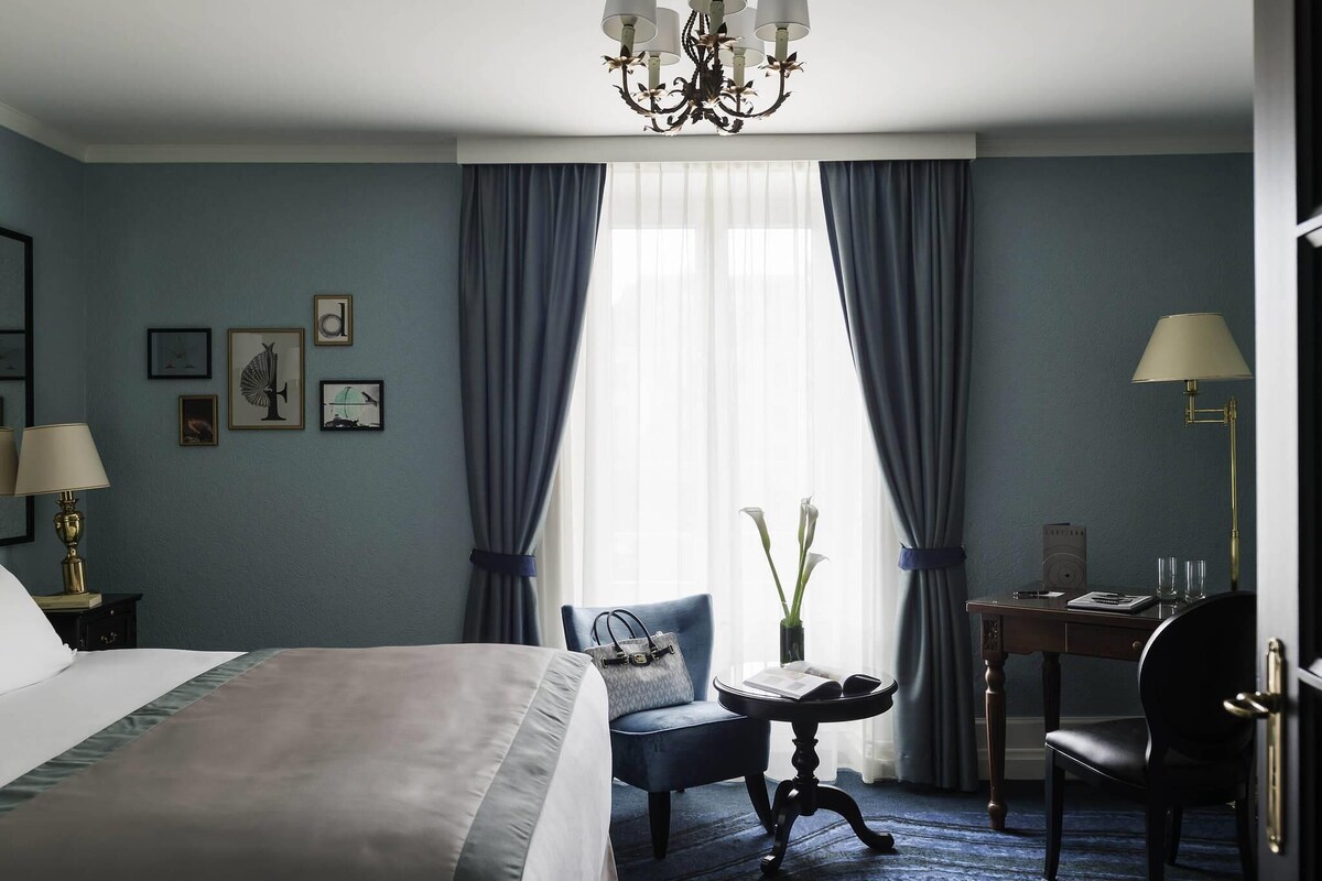 Et hotelværelse med seng, stol, skrivebord, lysekrone og gardiner.