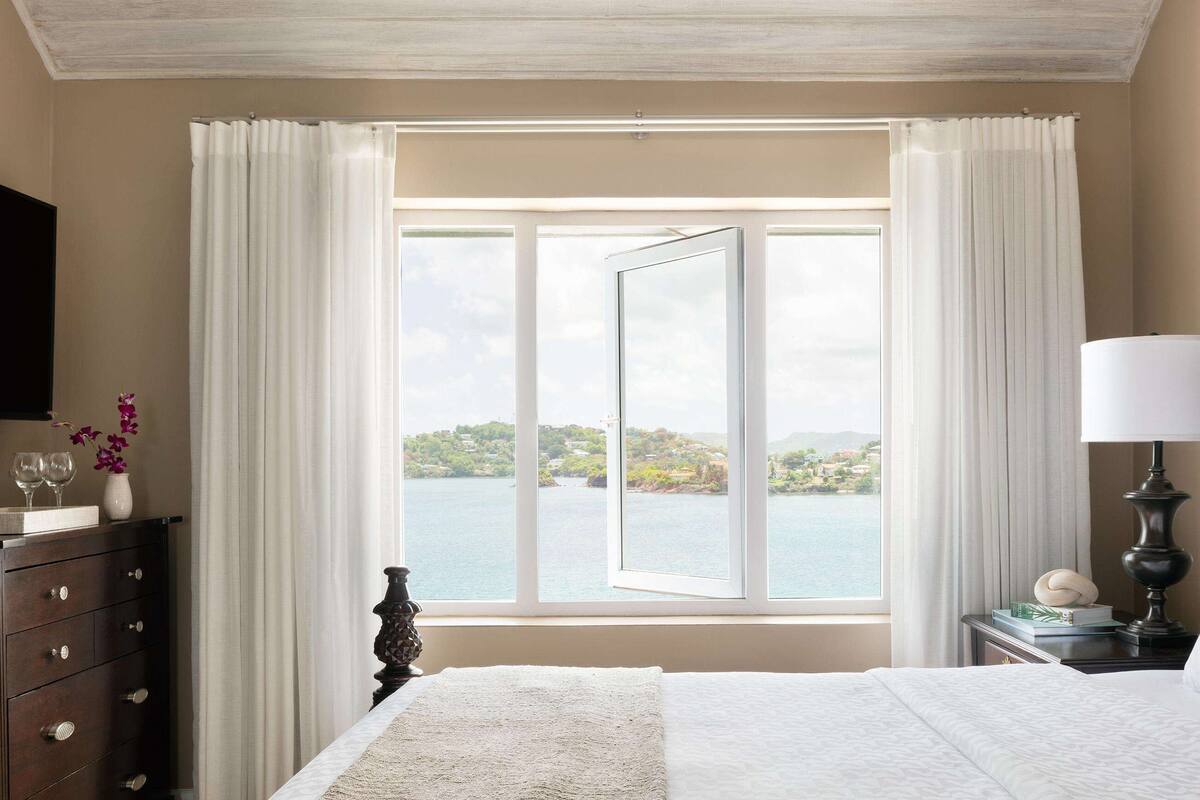 Une chambre avec une grande fenêtre, un lit, une table de chevet, une lampe, une commode et un téléviseur.