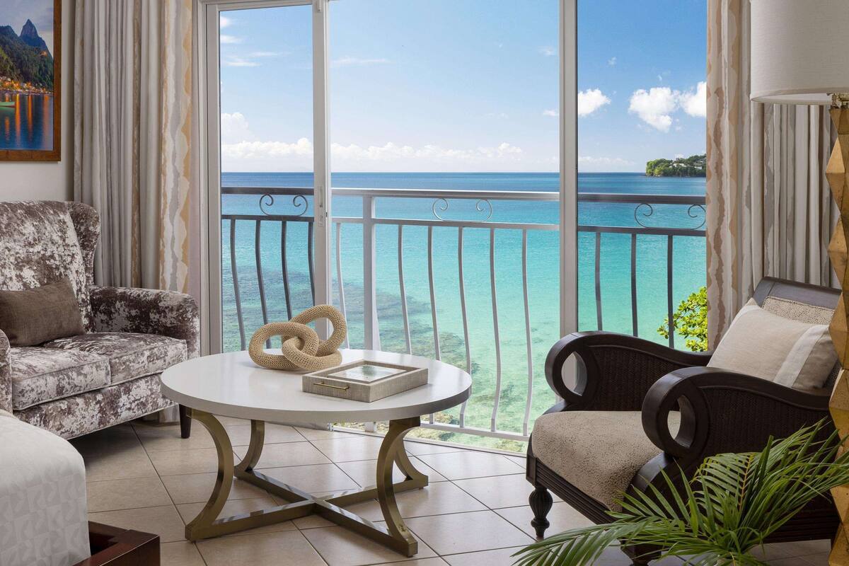 Une chambre dotée d’un balcon donnant sur une mer turquoise, et aménagée avec un canapé, un fauteuil et une table ronde.