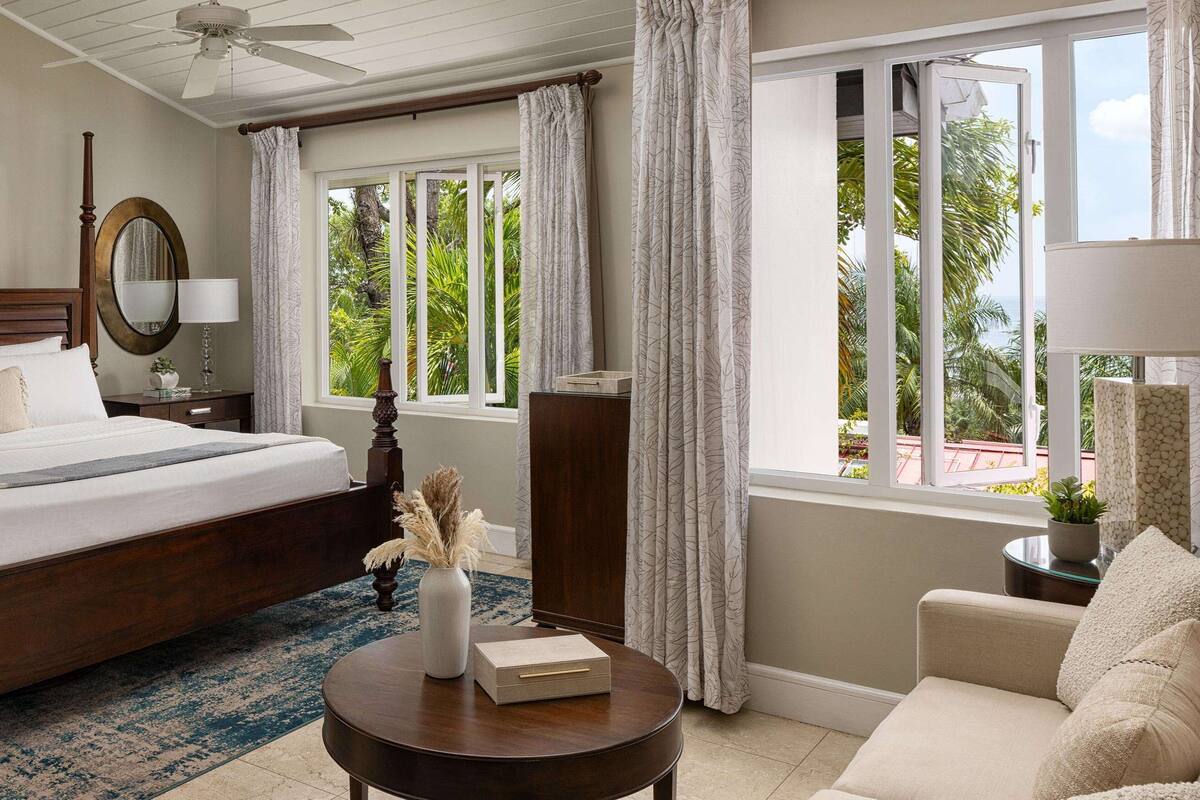 Une chambre d’hôtel avec un grand lit, une table ronde en bois, une chaise, un ventilateur de plafond et une vue sur l’extérieur.