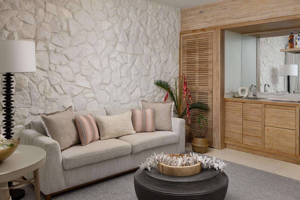 Un salon moderne avec un mur à l’aspect pierre, un canapé beige, une table basse ronde et un mini-réfrigérateur encastré doté d’une étagère pour les boissons.