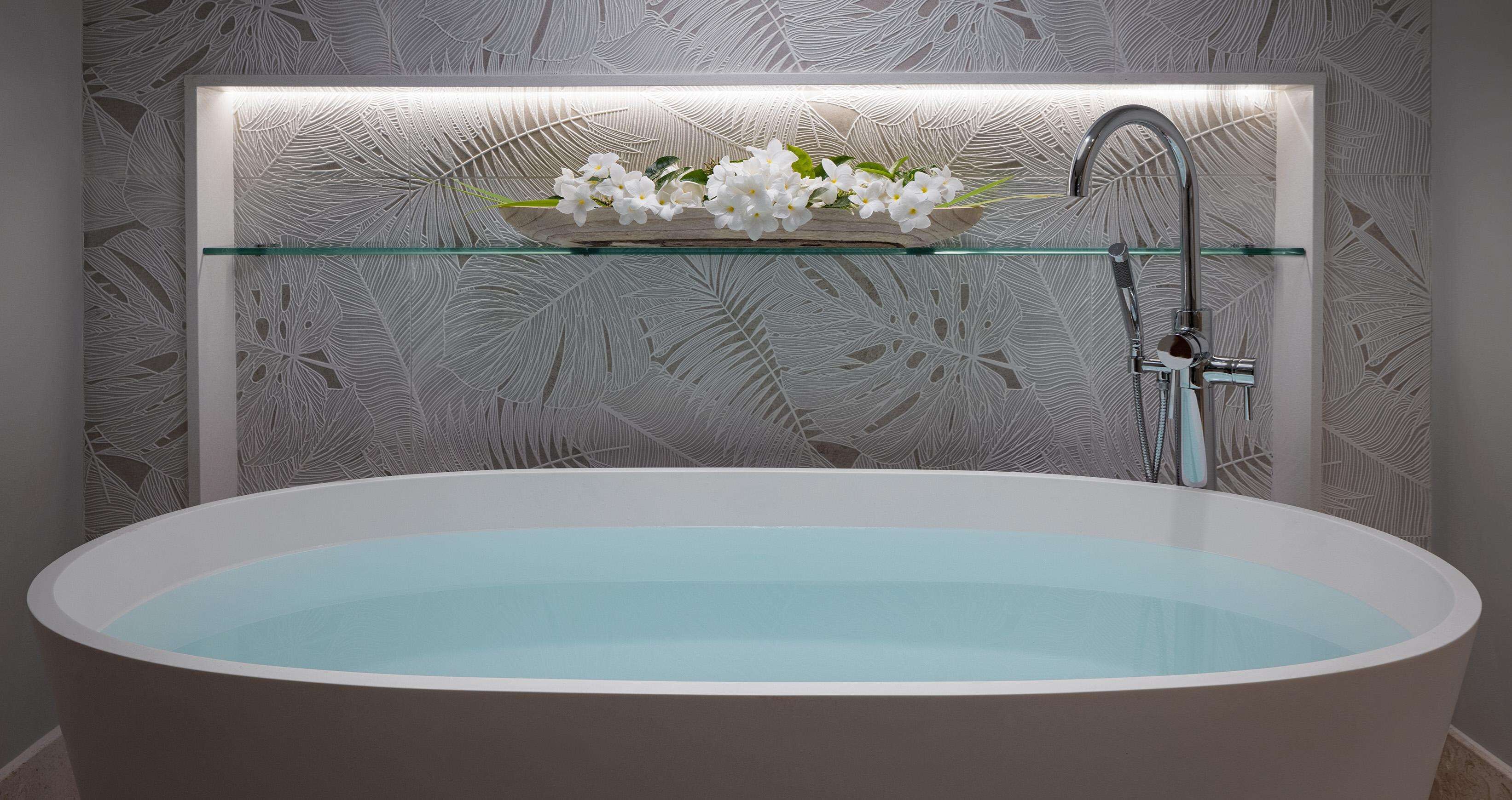 Een moderne badkamer met een vrijstaand bad, een glazen plank met bloemen, en een muur met een bladmotief.
