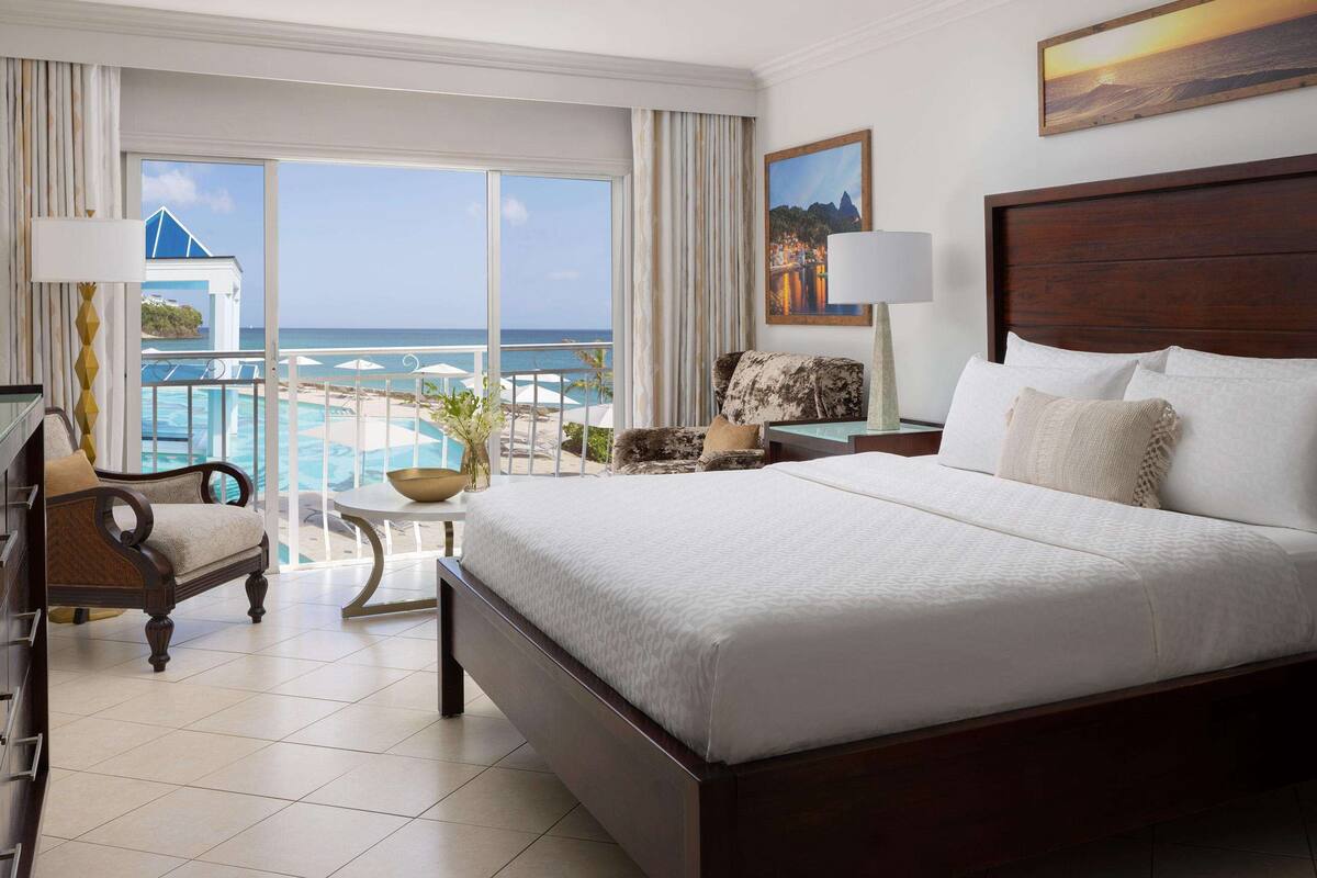 Une chambre d’hôtel avec un grand lit, une vue sur l’océan, un balcon avec une piscine et un fauteuil.