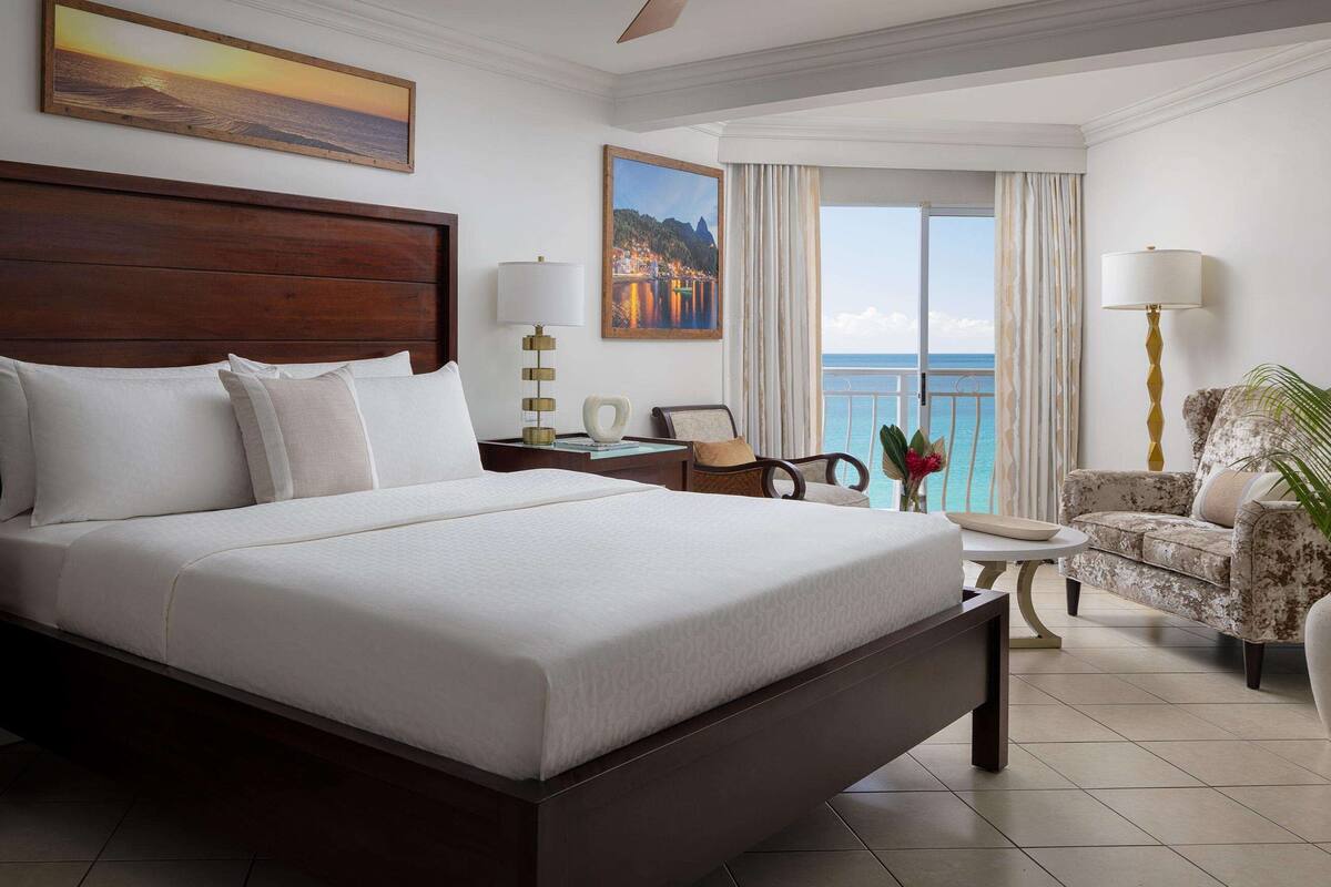 Une chambre d’hôtel avec un grand lit, une tête de lit en bois, une vue sur l’océan, une chaise et une table de chevet avec une lampe.