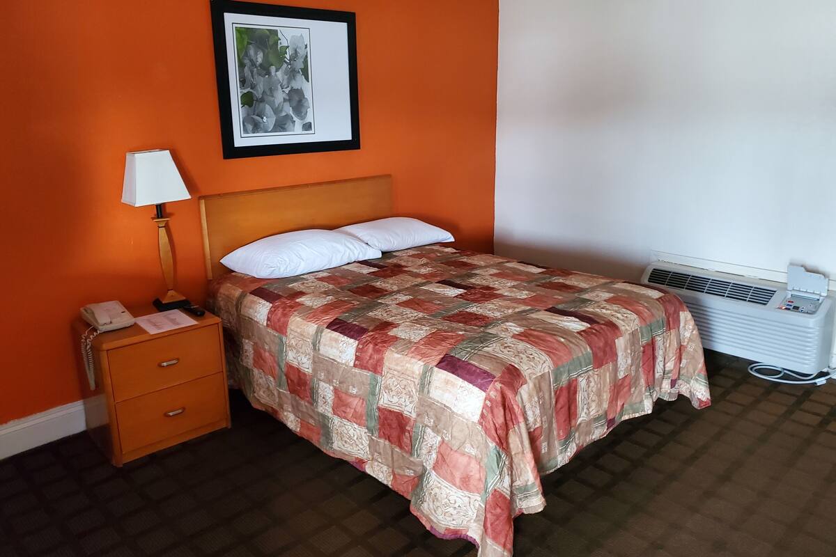 A hotel room with a bed, a nightstand with a lamp, a telephone, and a wall-mounted air conditioner.
