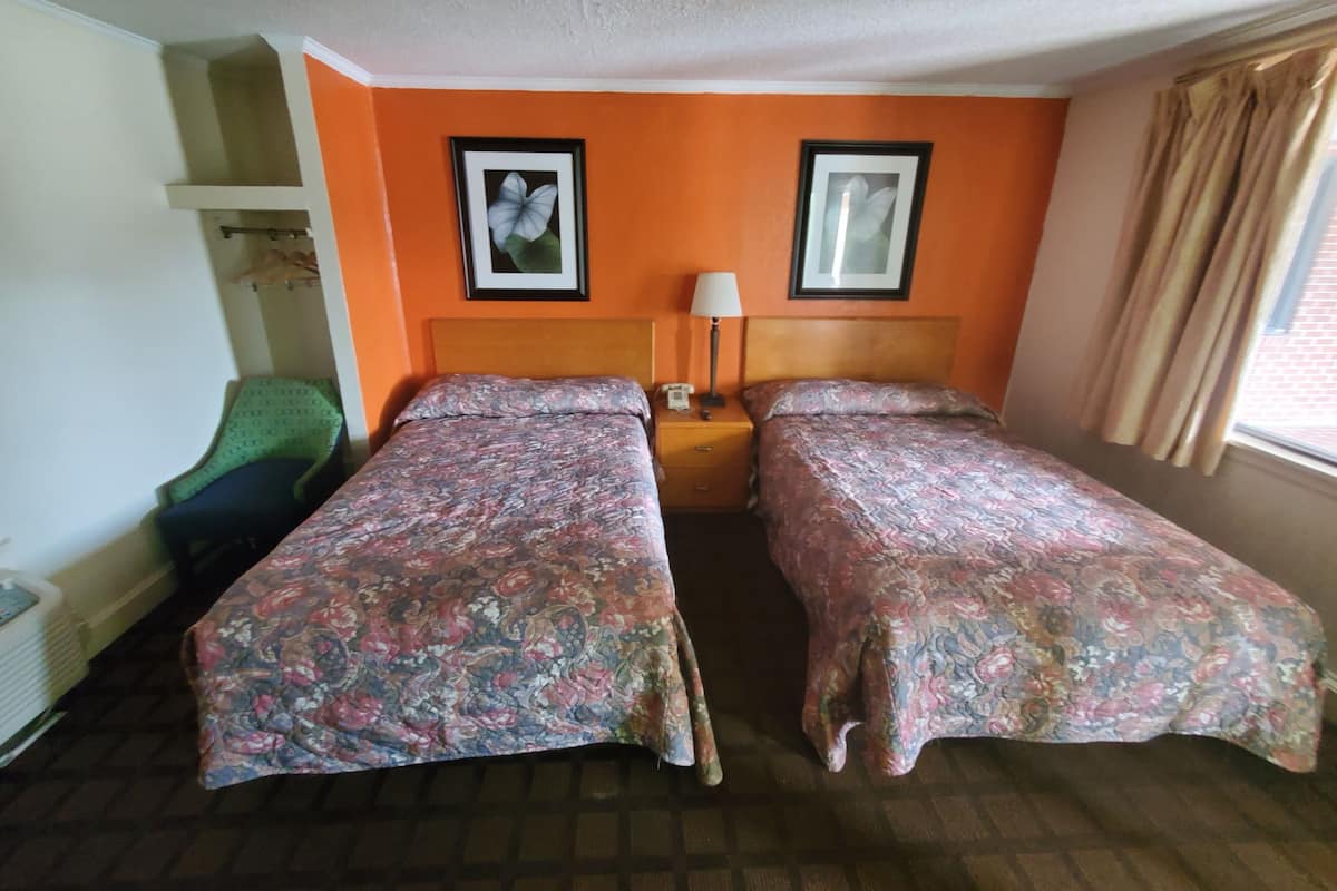 A hotel room with two single beds, a nightstand with a lamp, a chair, and a framed picture on the wall.