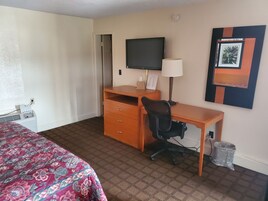 Habitación de hotel con una cama, un escritorio con silla, un televisor, una lámpara y un cesto de basura.
