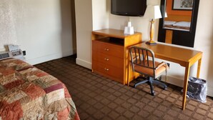 A hotel room with a bed, a desk, a chair, a television, a lamp, and a trash bin.