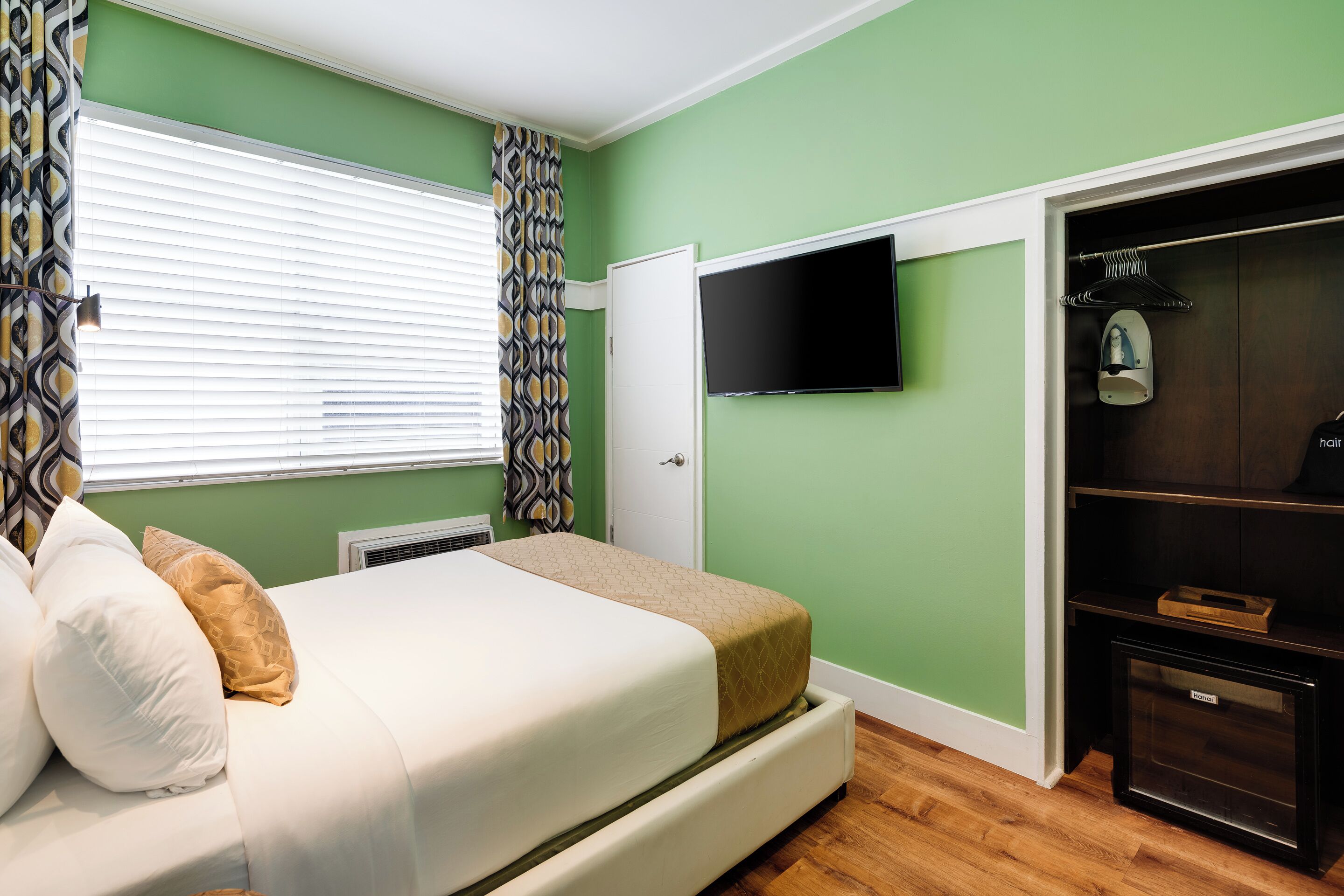Een slaapkamer met een bed, een aan de muur bevestigde televisie, een kledingkast en een raam met jaloezieën.