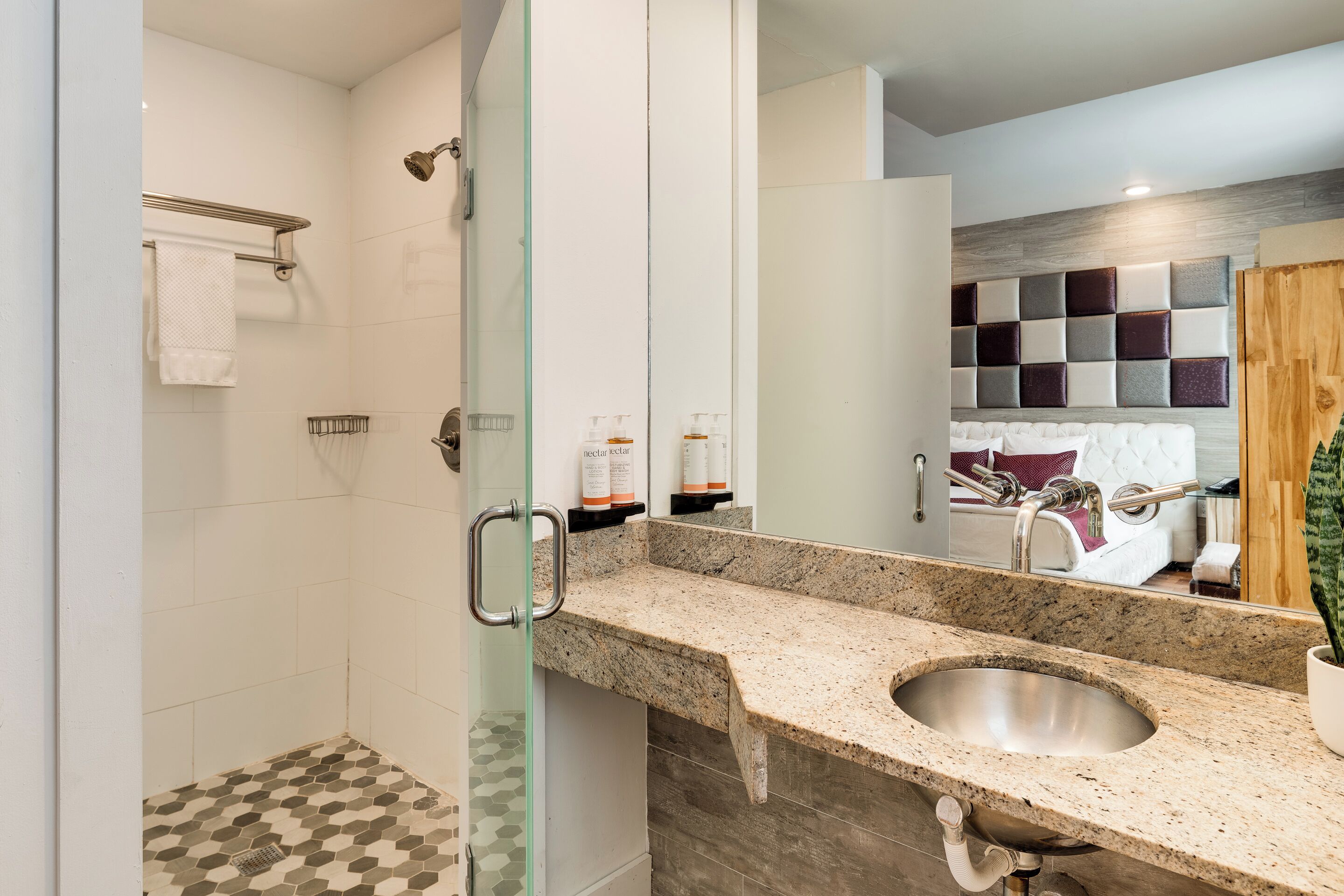 A modern bathroom with a glass-enclosed shower, a granite countertop sink, and a mirror reflecting a bedroom with a bed and a decorative wall panel.