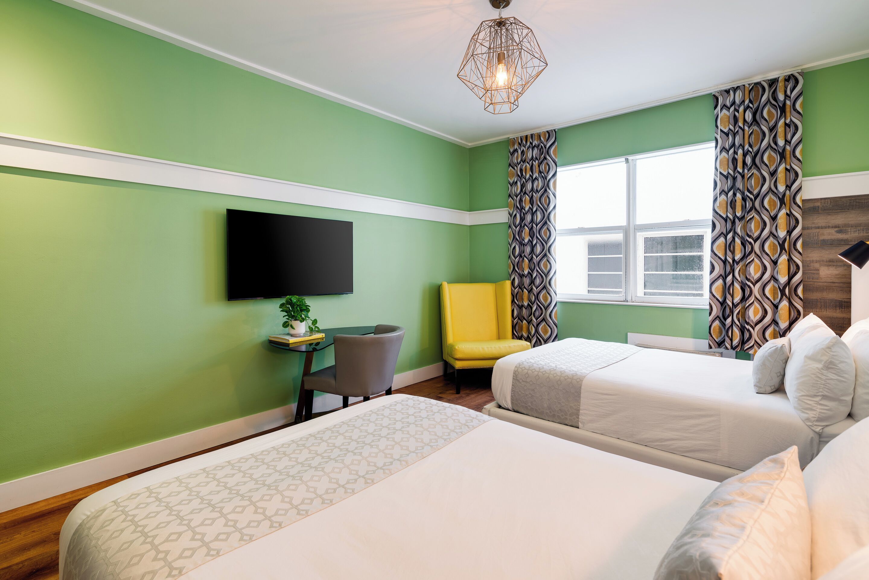 A hotel room with two beds, a desk with a chair, a flat-screen TV, and a window with patterned curtains.