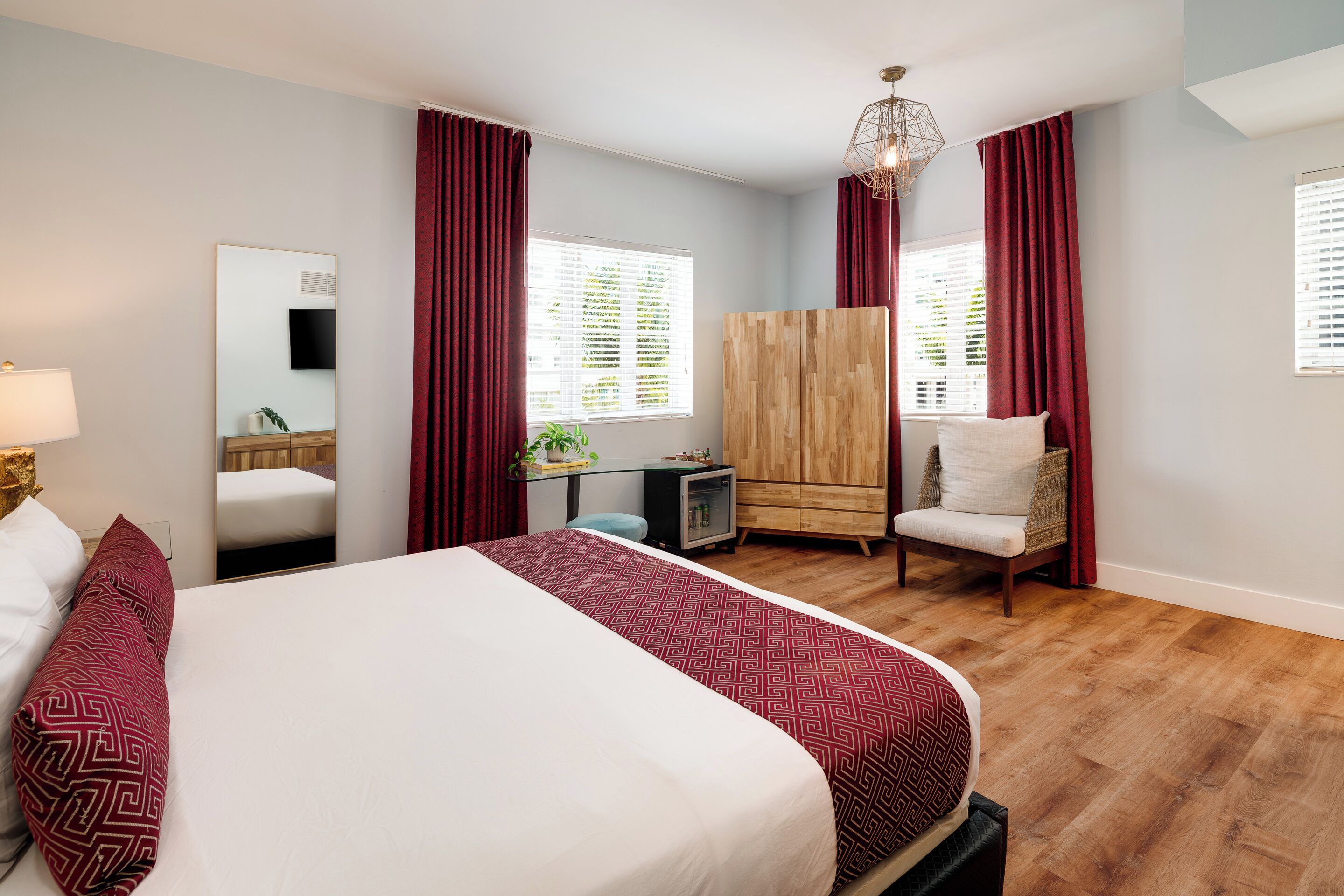 A bedroom with a large bed, a wooden wardrobe, a chair, a desk with a lamp, and a mirror.