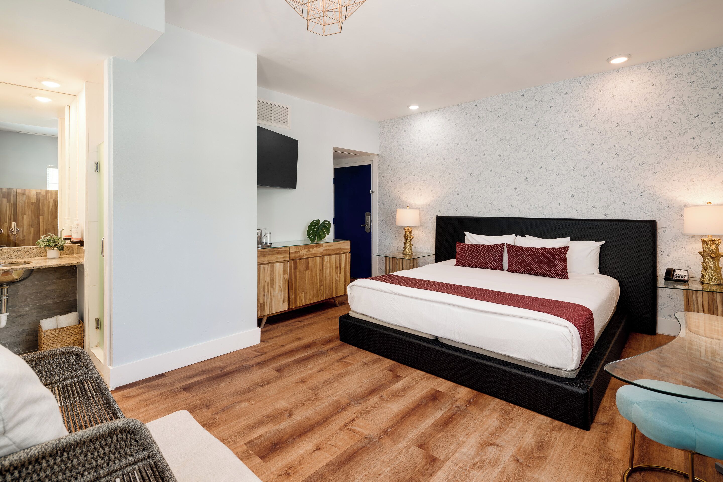 Una habitación de hotel moderna con una cama grande, piso de madera, un pequeño rincón de descanso y un baño a la vista a través de una puerta abierta.