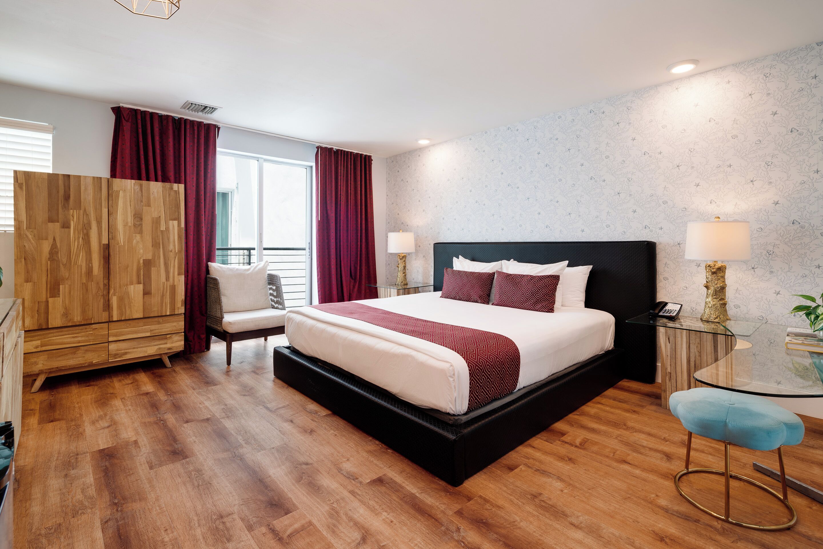 A modern hotel room with a large bed, a wooden wardrobe, a small table with a lamp, and a window with curtains.