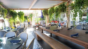 Un café en plein air avec des tables et des chaises, abrité par une pergola et entouré de plantes en pot.
