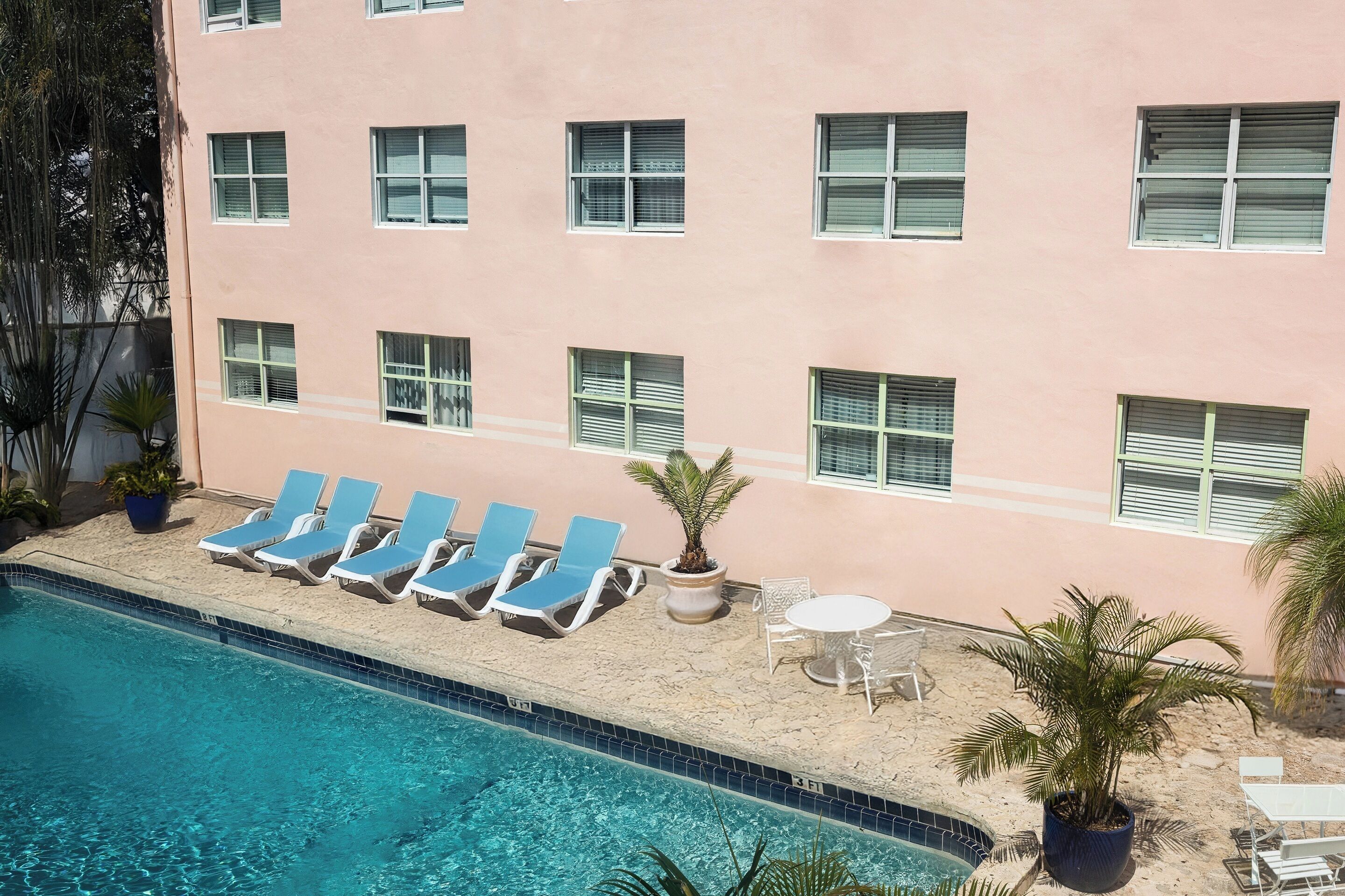 A hotel pool area with lounge chairs and a small table, surrounded by potted plants.