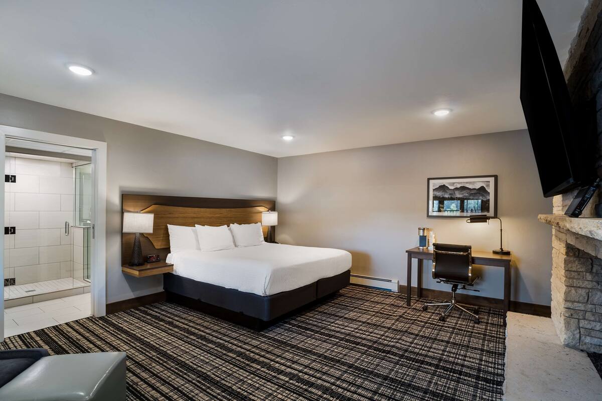 A hotel room with a large bed, a bedside table with a lamp, a small desk with a chair, a fireplace, and a framed picture on the wall.