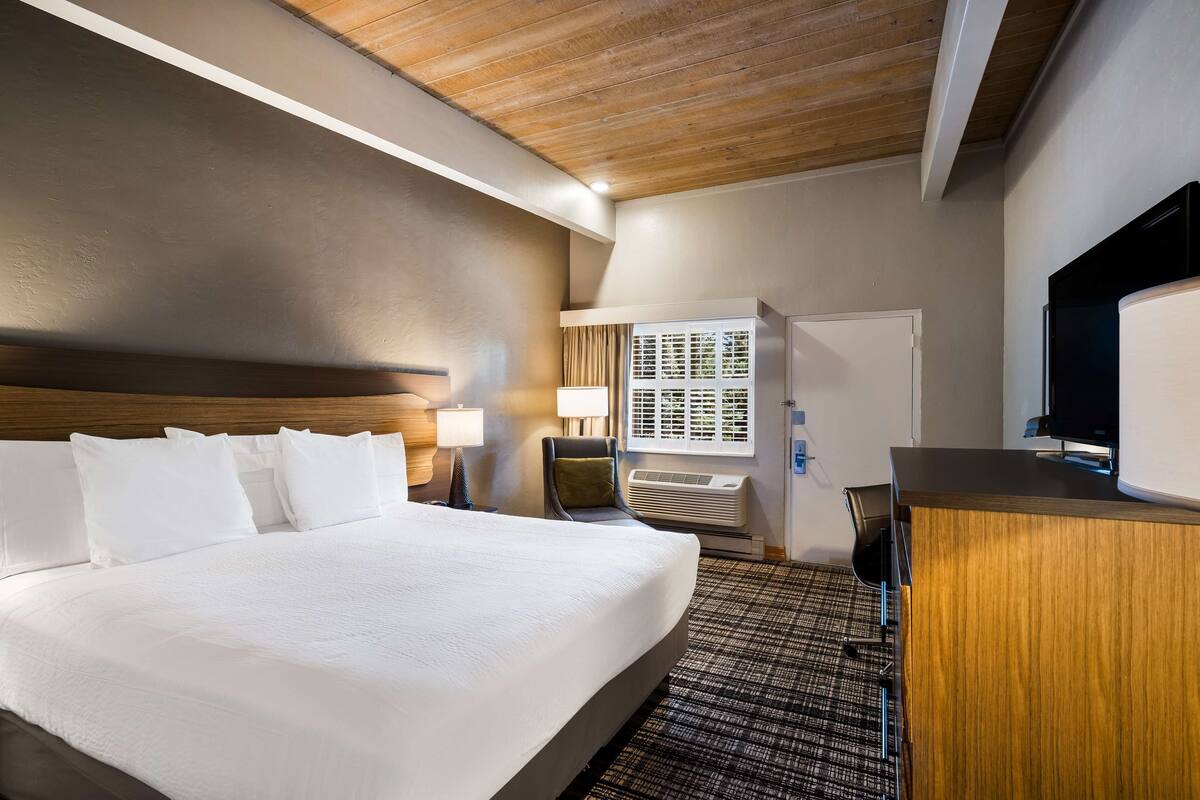 A hotel room with a large bed, a wooden ceiling, a desk with a TV, and a window with blinds.