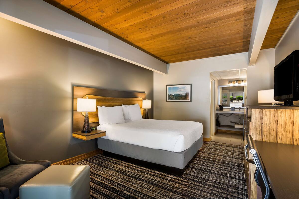 A hotel room with a bed, bedside lamps, a TV, a desk, and a view of the hallway and another room.