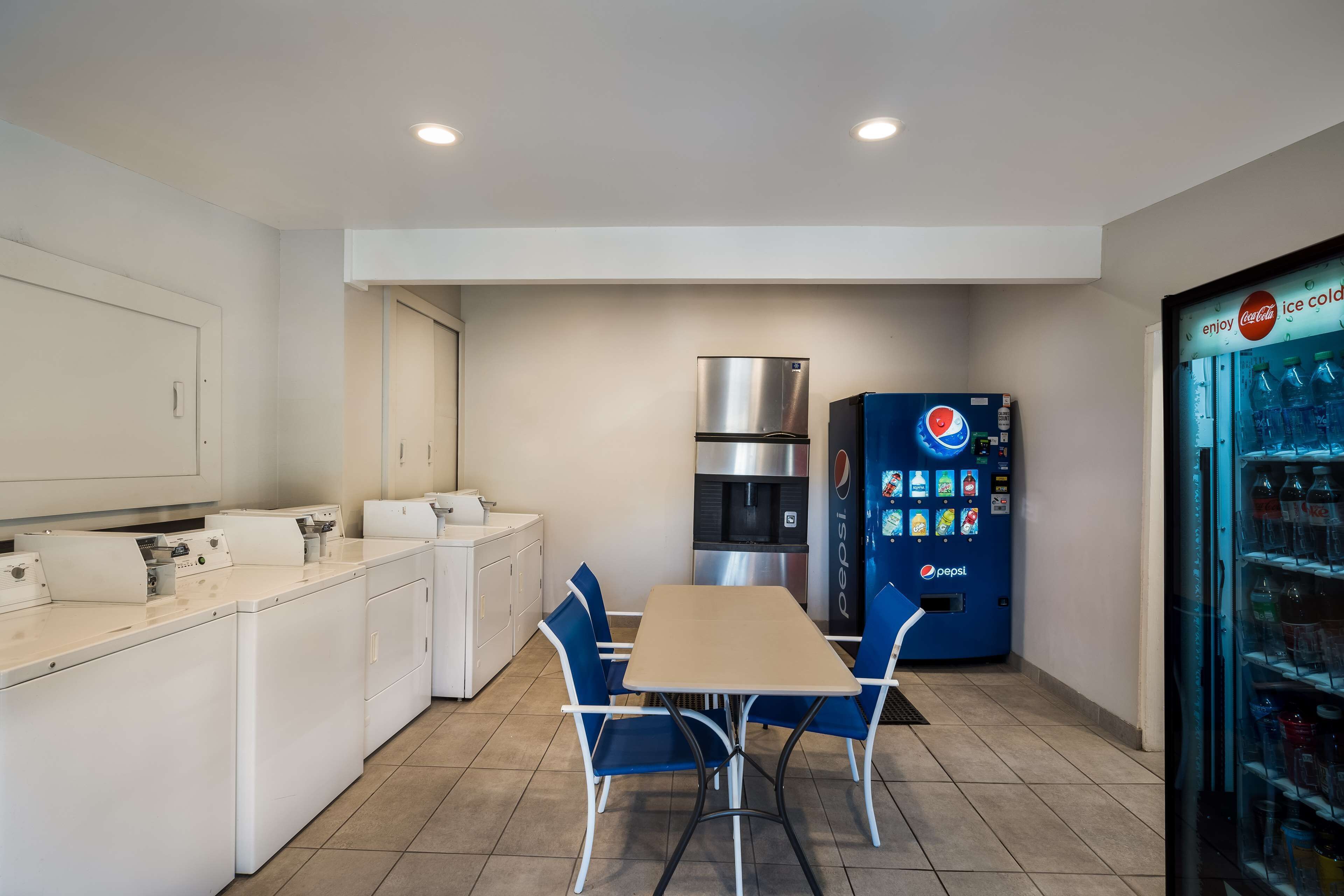 Una cocina compacta con lavadora, refrigerador, una mesa con sillas y una máquina expendedora de Pepsi.