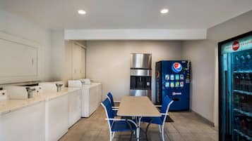 Una cocina compacta con lavadora, refrigerador, una mesa con sillas y una máquina expendedora de Pepsi.