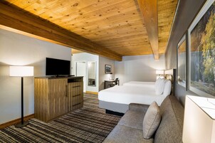 Quarto de hotel com teto de madeira, uma cama grande, um sofá, uma televisão sobre um móvel de madeira e um quadro na parede.
