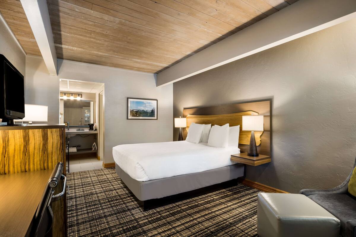 A hotel room with a bed, a desk, a chair, a TV, and a bathroom visible through an open door.
