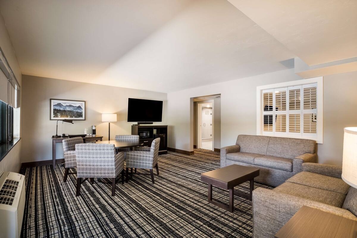 A hotel room with a flat-screen TV, a desk with a lamp, two patterned chairs, a sofa, and a large window with blinds.