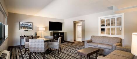 Habitación de hotel con televisión de pantalla plana, un escritorio con lámpara, dos sillones estampados, un sofá y un ventanal con persianas.