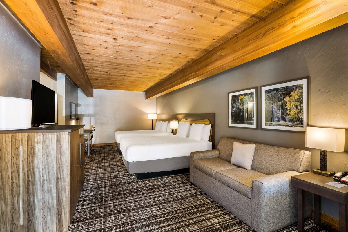 A hotel room with a wooden ceiling, a large bed, a sofa, a desk with a lamp, and two framed pictures on the wall.