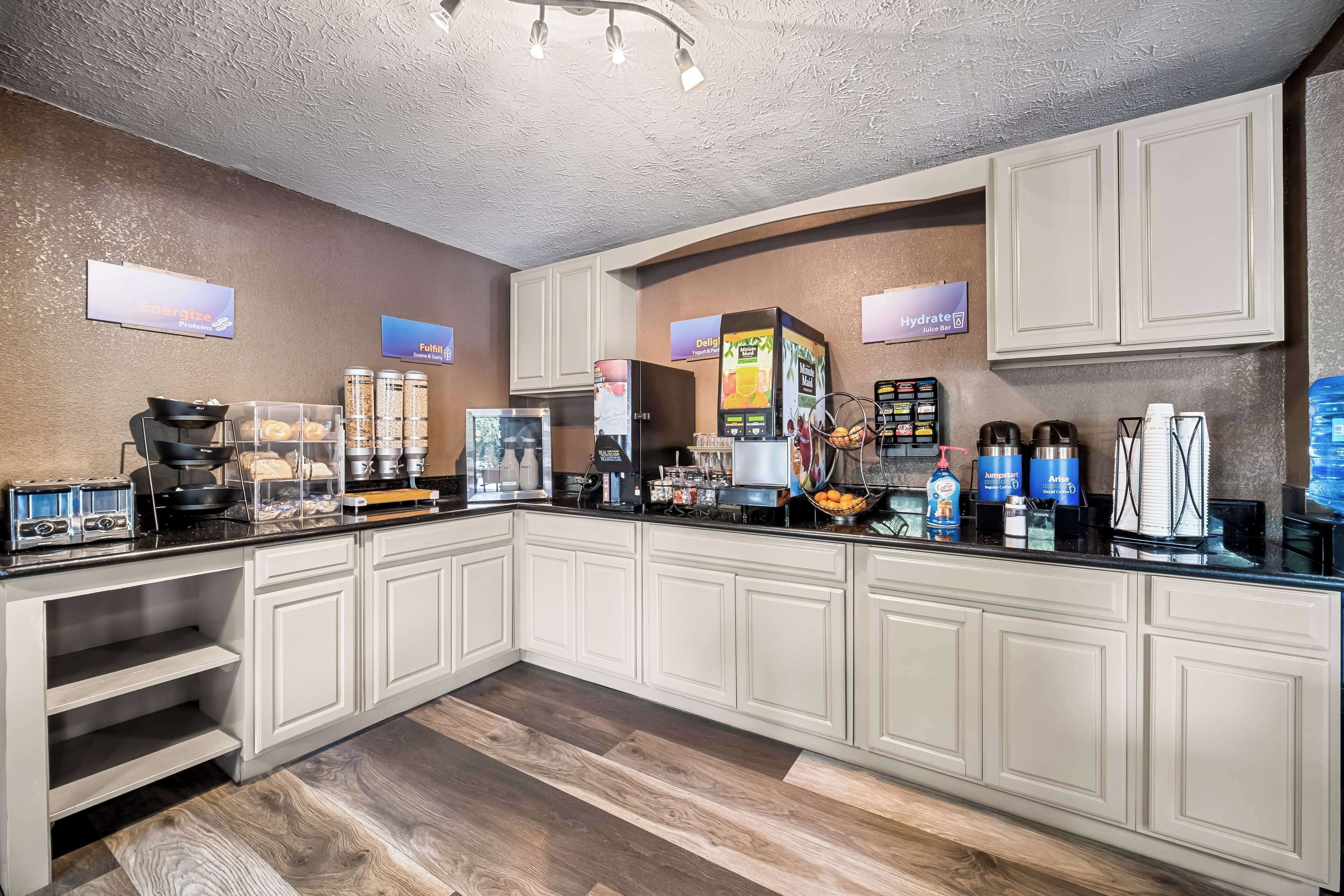 A modern kitchen with white cabinets, a black countertop, and various appliances including a coffee machine, a kettle, and a microwave. There are also signs for 'Energy' and 'Hydrate' on the wall.