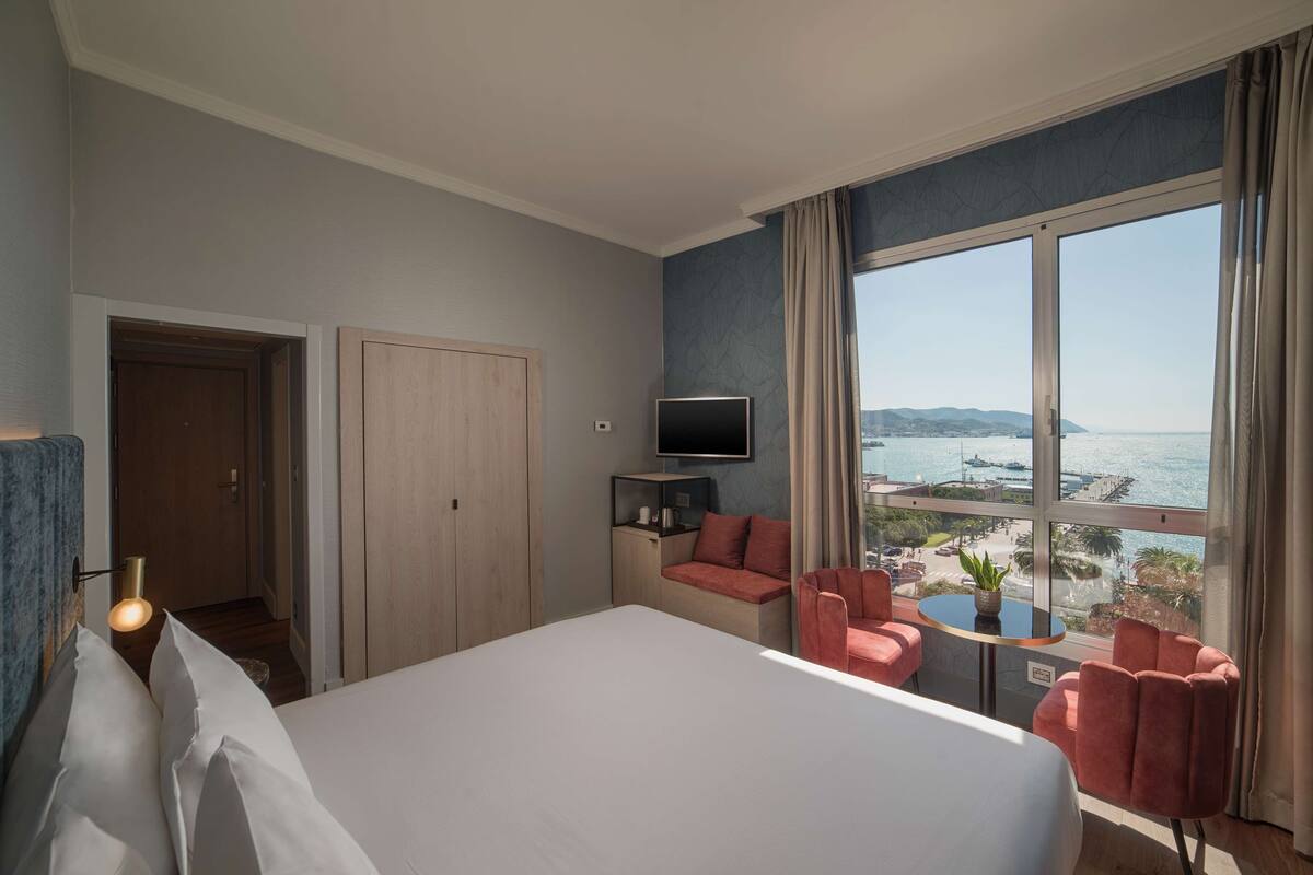 Une chambre d’hôtel avec un grand lit, un coin salon avec des fauteuils, une télévision et une vue sur la mer.