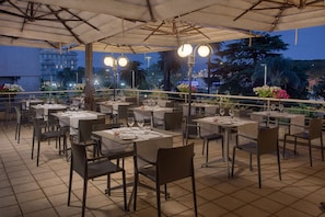 Un espacio exterior para comer con mesas y sillas, cubierto por un techo, y vistas a un paisaje urbano.