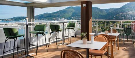 A deck with outdoor seating, a view of the sea, and mountains in the background.