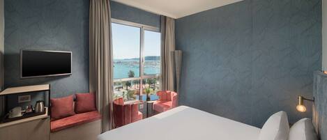 Habitación de hotel con cama, televisión, escritorio con hervidor, una mesita y vistas al mar.