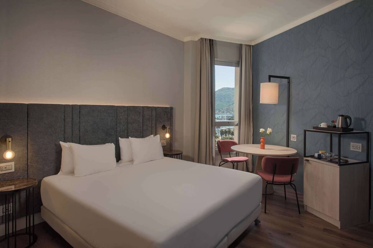 Une chambre d’hôtel avec un grand lit, une petite table ronde avec des chaises, un bureau avec une lampe et une vue sur l’extérieur.