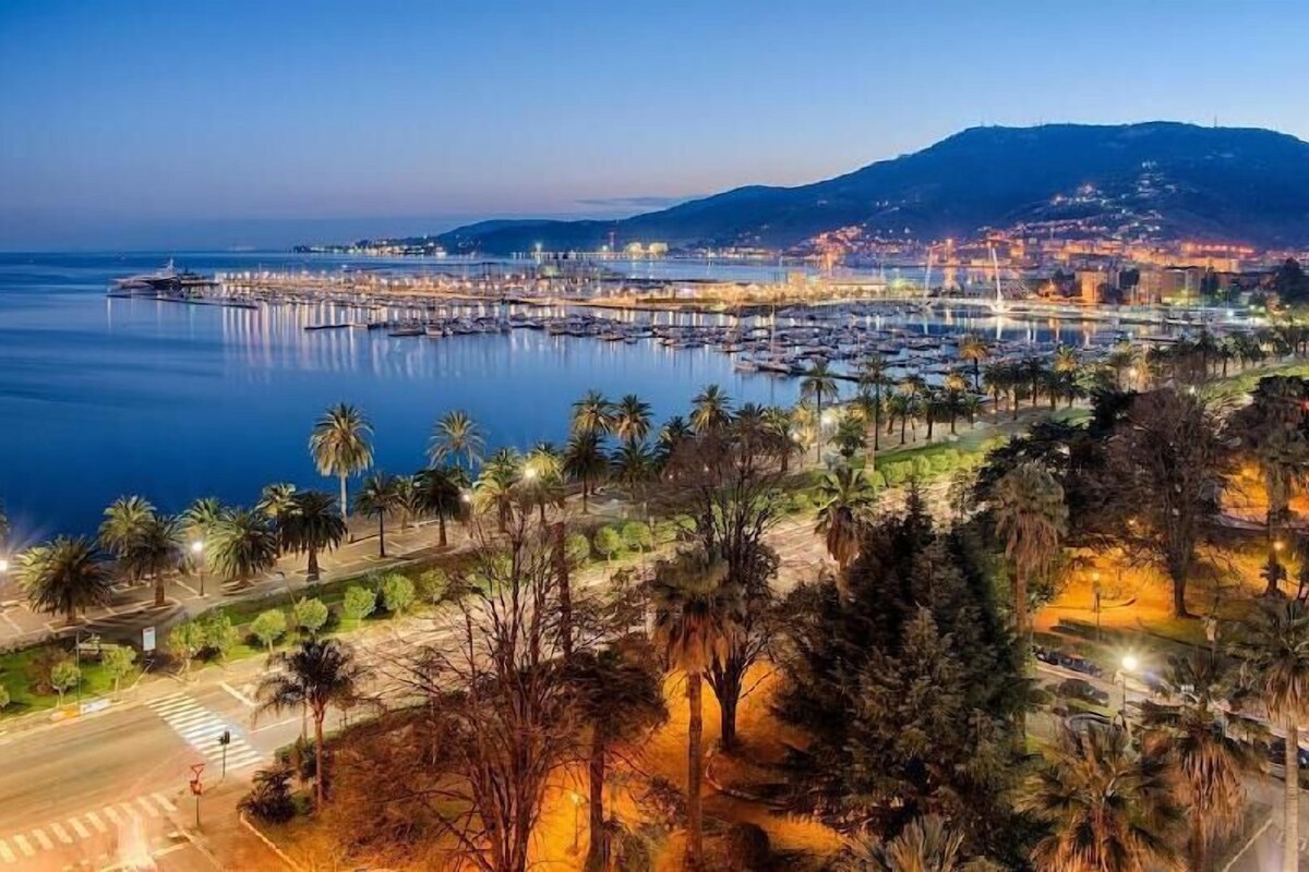 Un paysage urbain côtier avec un port de plaisance, une promenade bordée de palmiers et une montagne en arrière-plan.