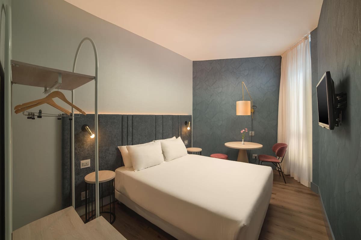 Une chambre d’hôtel avec un grand lit, deux petites tables rondes, un téléviseur fixé au mur et un porte-manteau.