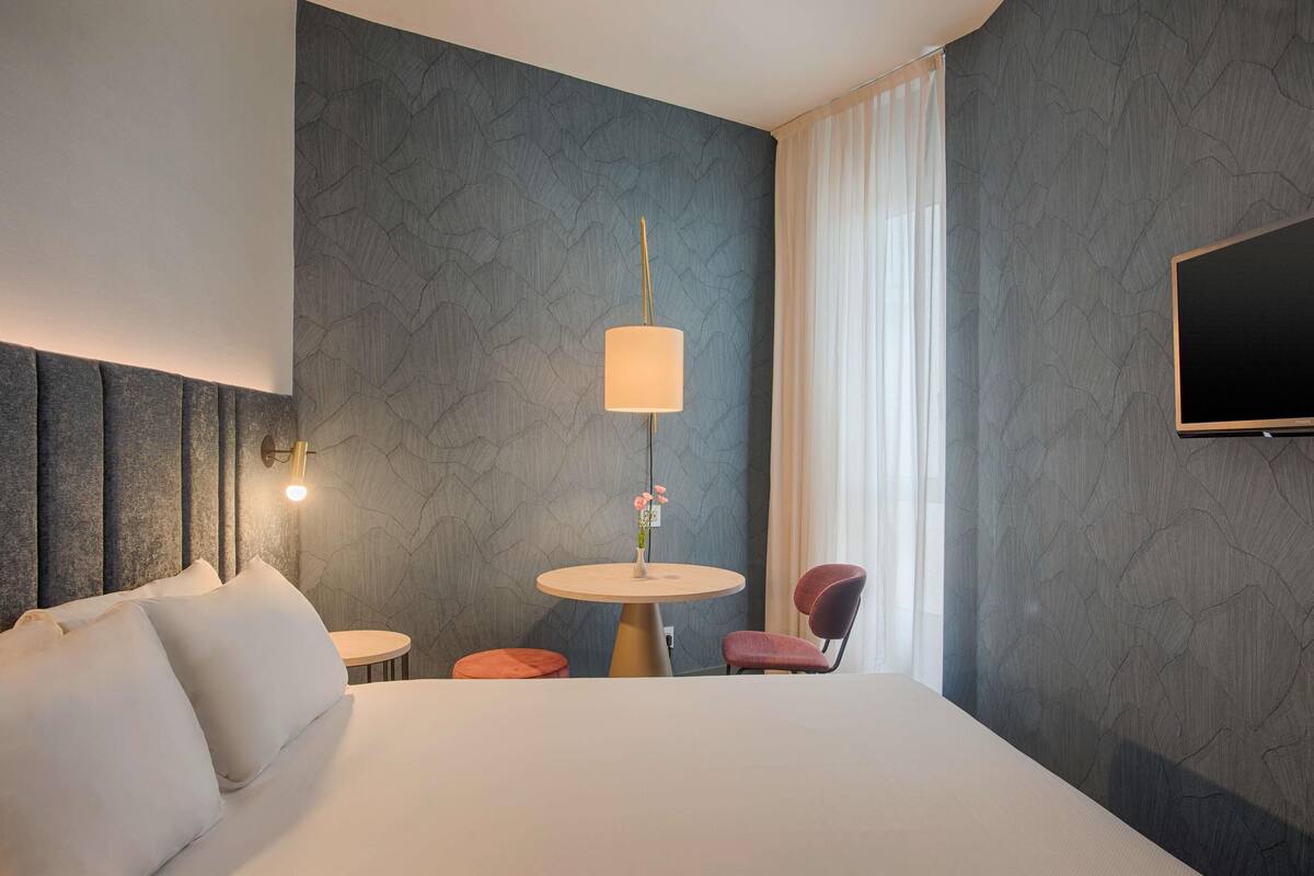 Une chambre d’hôtel avec un lit, une table ronde avec une lampe, une chaise et un téléviseur fixé au mur.